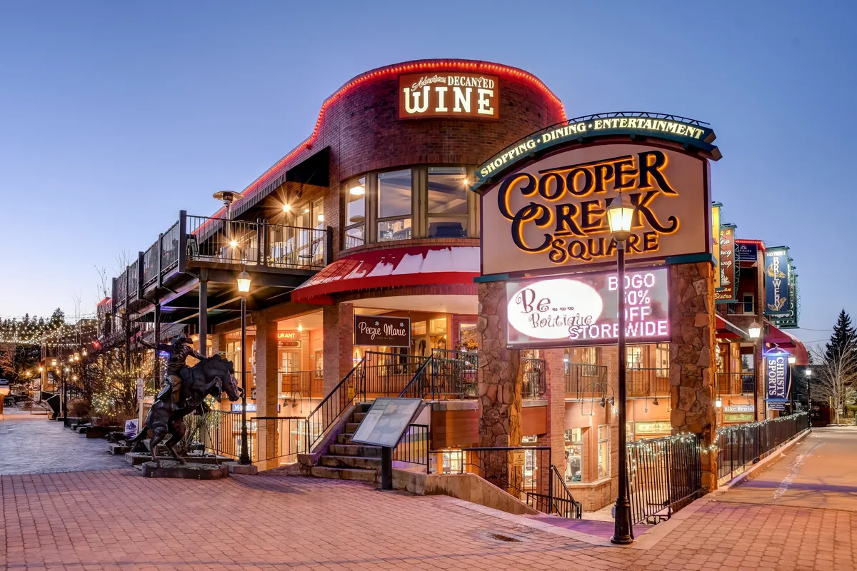 An inviting exterior of Cooper Creek Square with its illuminated sign, wine shop, decorative string lights, and public art sculpture. People gather for shopping, dining, and live entertainment, suggesting a lively downtown Winter Park scene.