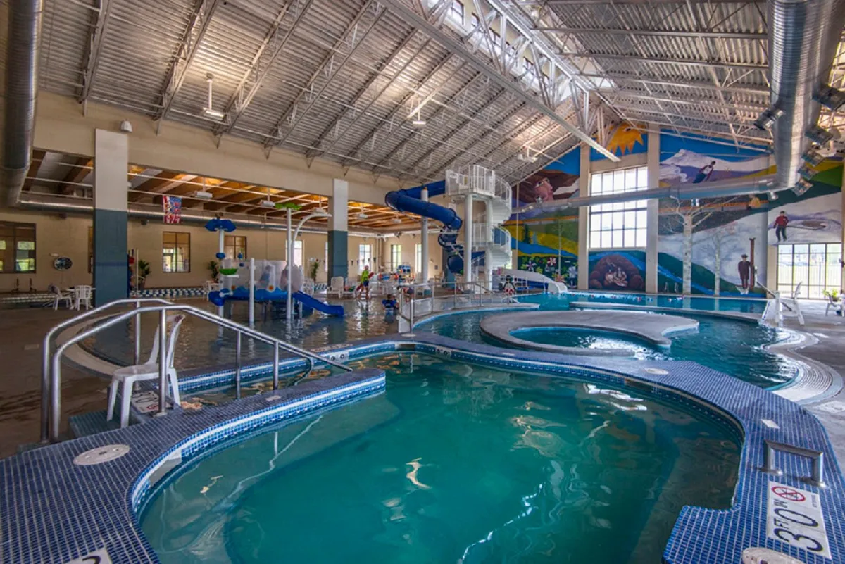 A bright and bustling indoor recreation center scene featuring colorful water slides, splash areas, swimming pools, and families relaxing on lounge chairs. Kids and adults are shown playing and bonding, illustrating the fun and community vibe of the Grand Park Rec Center.