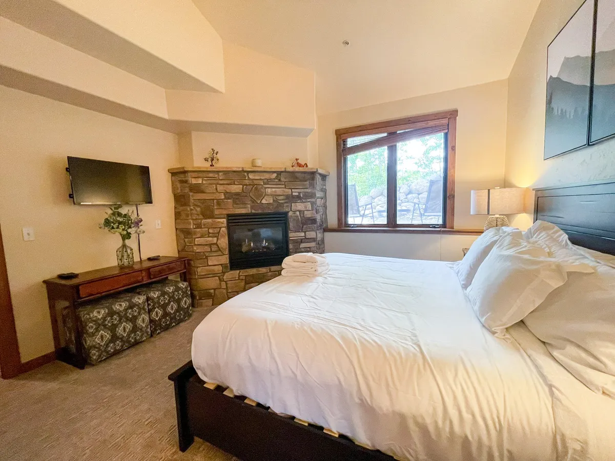 This image features a king-size bed facing a striking stone fireplace, with a large window allowing an abundance of natural light and scenic views. The room includes a TV and a decorative plant, giving it a cozy yet upscale atmosphere ideal for unwinding after a day of adventure.