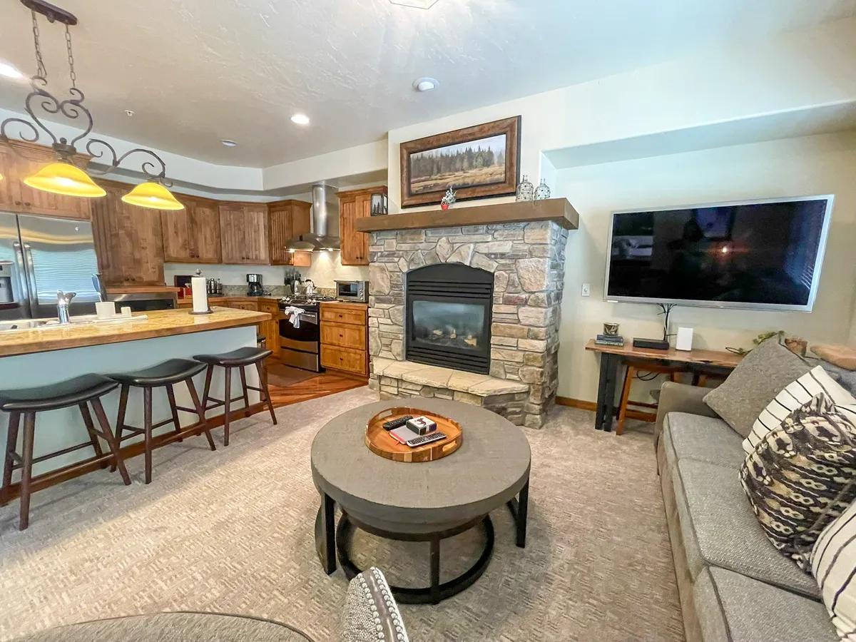 A warm, welcoming interior scene that highlights a cozy living space with a roaring fireplace, a comfortable couch, a kitchen island, and a coffee table. The open-concept design and inviting atmosphere evoke togetherness, relaxation, and the comforts of home after a day outdoors.