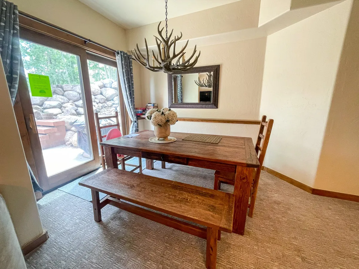 A warm, rustic dining area with a large wooden dining table, an antler chandelier above, a large mirror reflecting natural light, and a sliding glass door offering a glimpse of the outdoors. The table is set for family meals, and the ambiance feels cozy and festive.