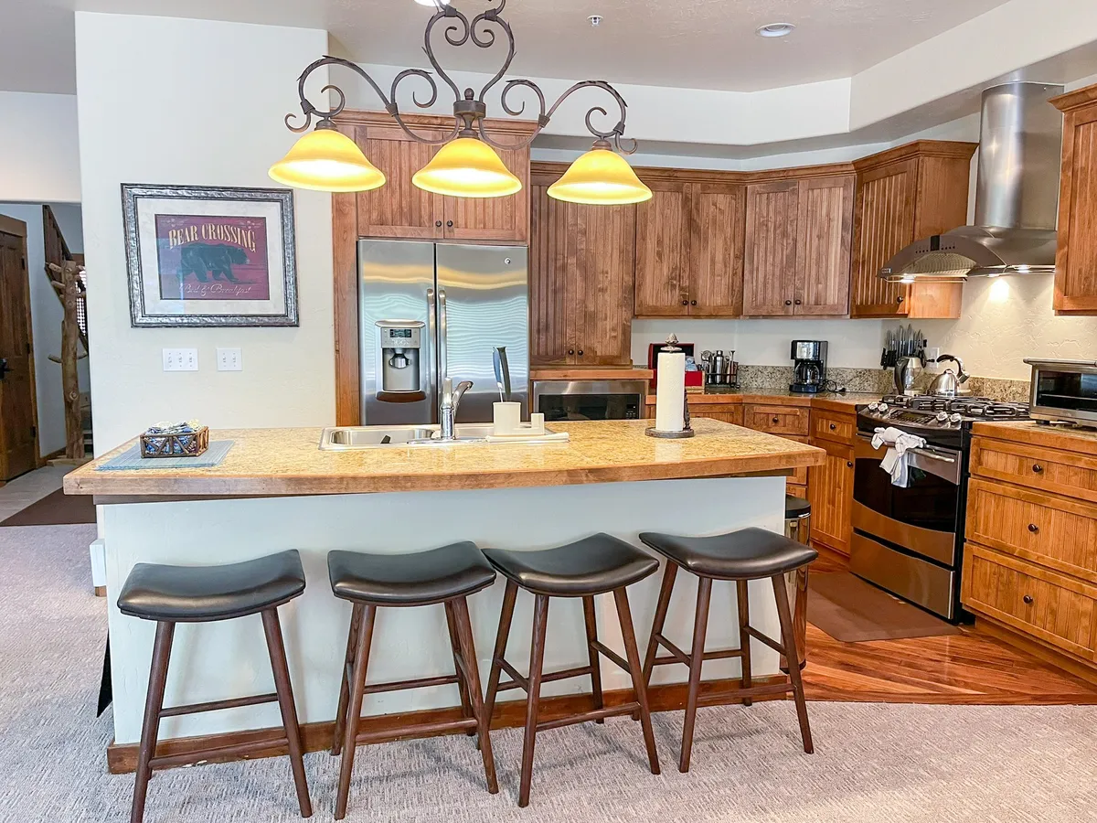 A contemporary chef’s kitchen with a spacious island and bar stools, a striking chandelier, wood cabinetry, and stainless steel appliances. The kitchen is designed for gourmet cooking, entertaining, and informal gatherings.