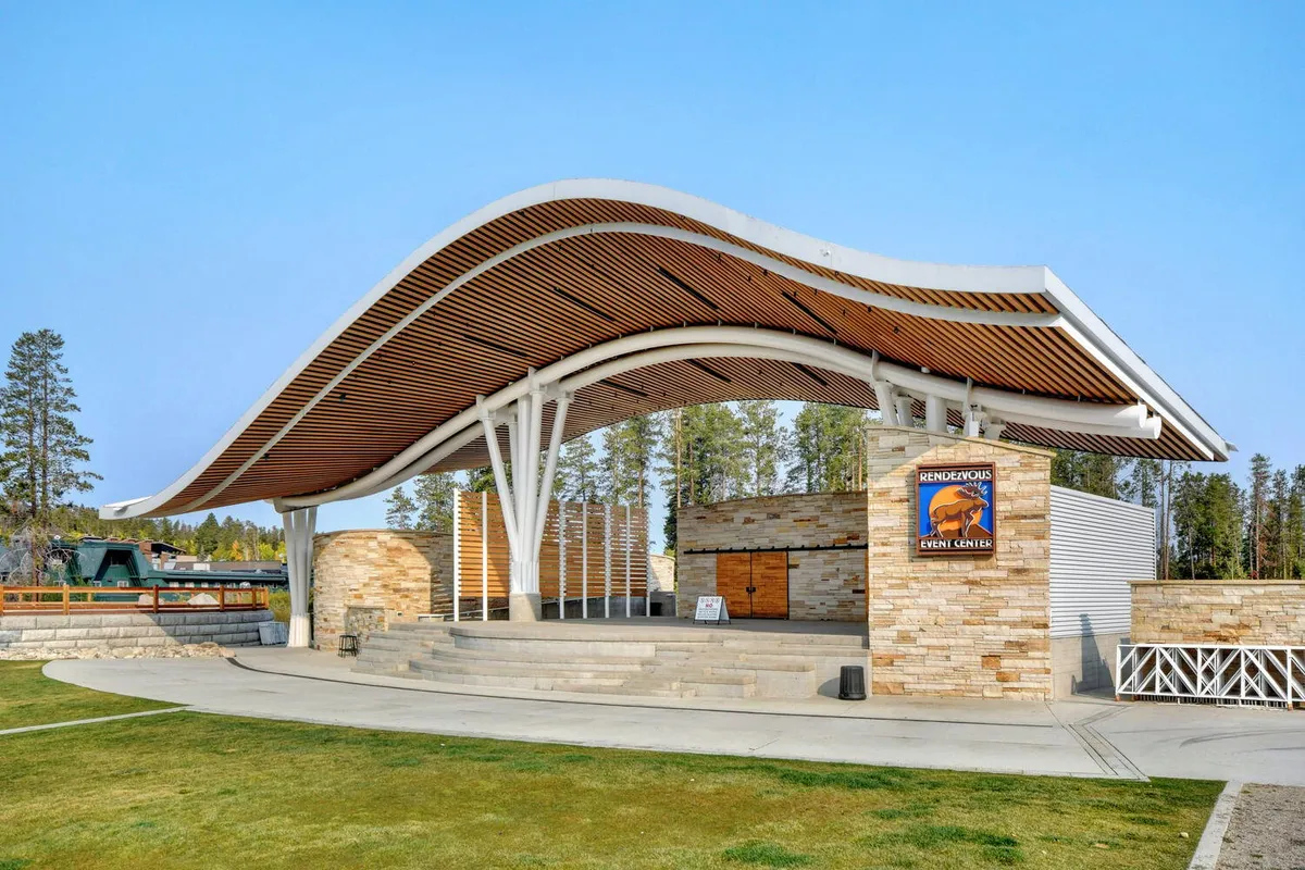 An exterior scene featuring a striking curved roof structure with a stone façade and an open stage area. The setting is ideal for outdoor concerts, community gatherings, and art showcases, with expansive space that feels connected to the local environment and encourages community interaction.
