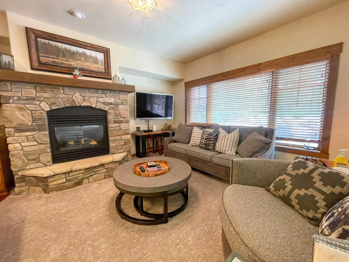 A spacious open-concept living room featuring a prominent stone fireplace, a circular coffee table, a comfortable couch, and expansive windows with blinds. The space exudes warmth and provides an inviting setting for families to relax, enjoy the fireplace, and take in the mountain views.