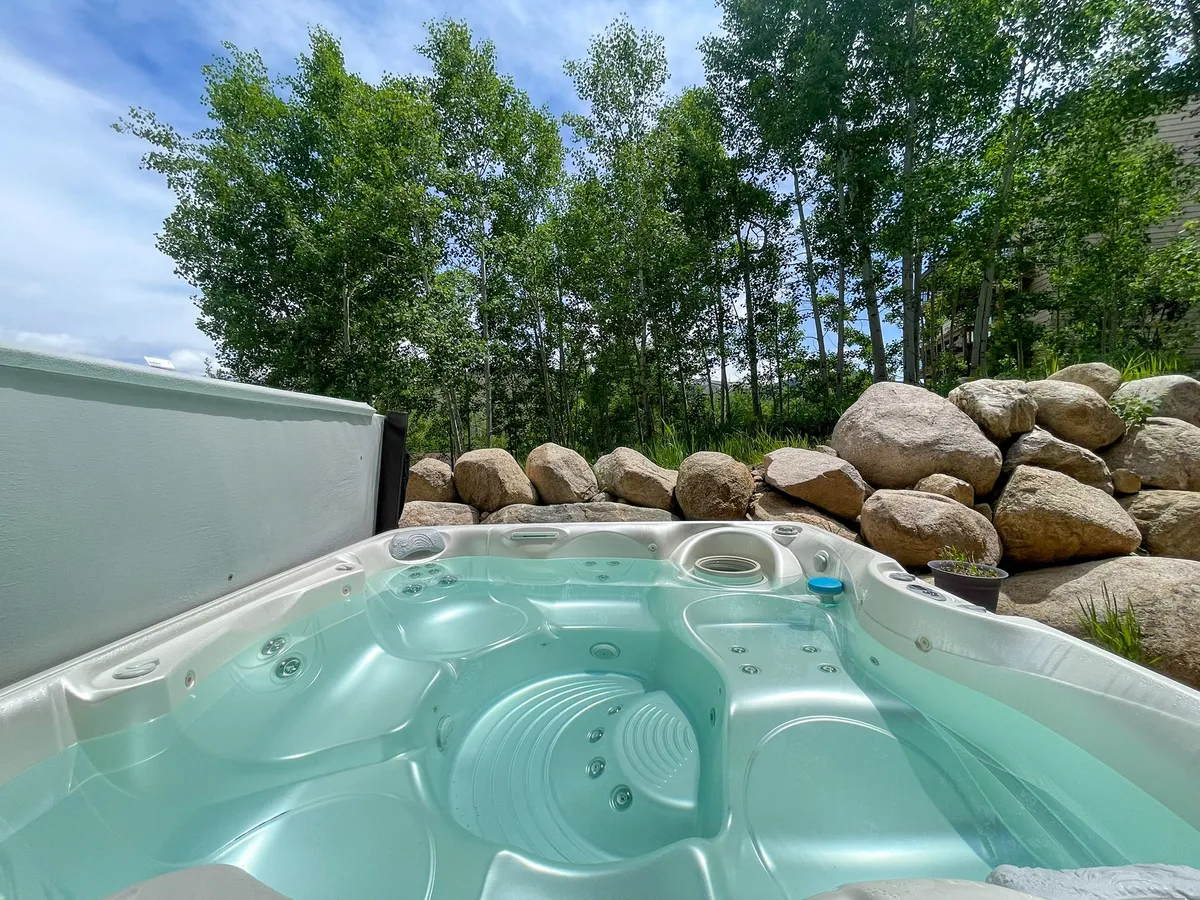 An inviting outdoor hot tub set among mature trees and natural rock formations, with people relaxing and socializing, suggesting both solitude and togetherness. The tranquil setting is ideal for stargazing or enjoying a morning coffee surrounded by nature.