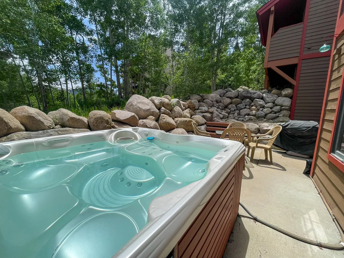 A spacious patio featuring a hot tub, patio furniture, and natural landscaping with rocks and trees. The scene suggests opportunities for stargazing at night, poolside drinks, and lively barbecues with friends or family.