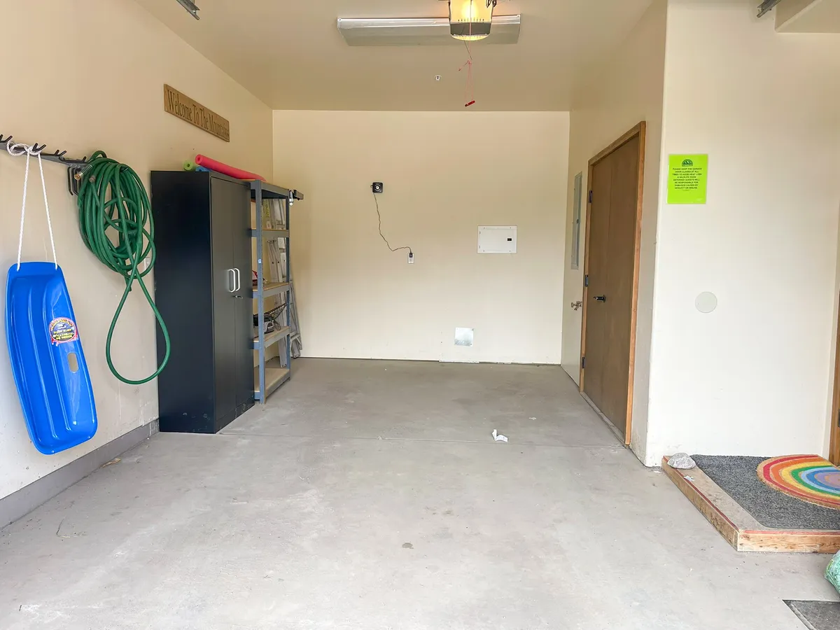 A bright and practical mudroom with organized storage, including a hanging garden hose, black shelving, a colorful sled, and a rainbow doormat. The space feels welcoming and functional, ideal for storing outdoor adventure gear.