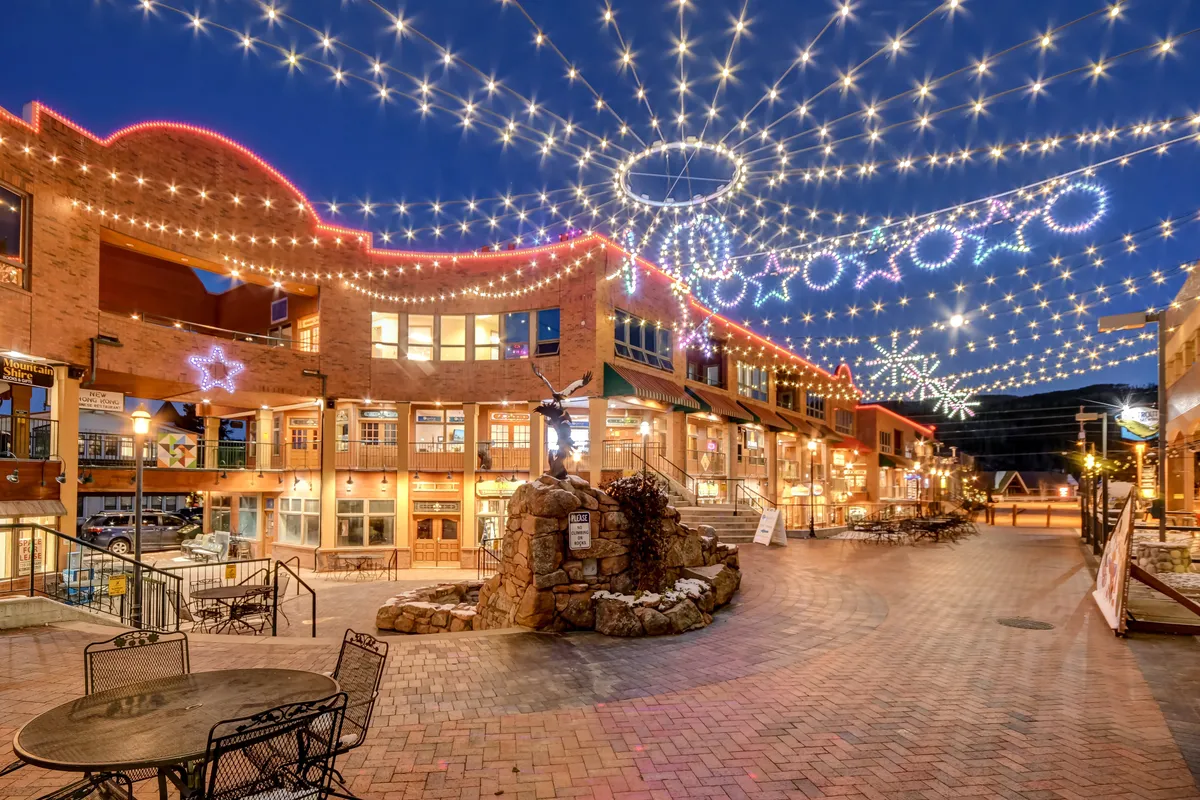 A bustling downtown street scene in Winter Park, with decorative lights strung above brick pathways, inviting storefronts, and a charming fountain. People are seen dining, shopping, and strolling, creating a festive, welcoming atmosphere that highlights the local community's vibrancy.